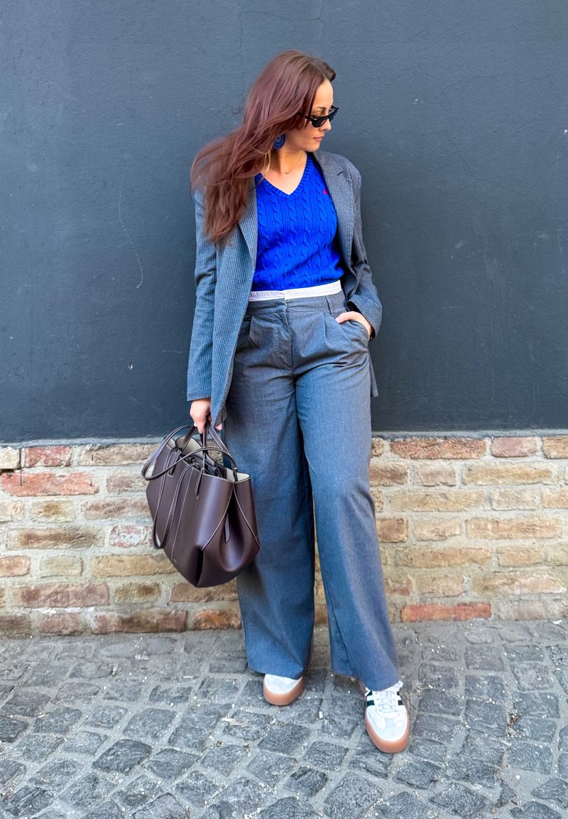 Femme aux longs cheveux bruns portant des lunettes de soleil, un pull bleu, un blazer gris et un pantalon, des baskets blanches, tenant un sac à main marron, debout dans une rue pavée.