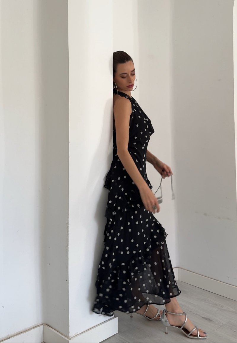 Femme en robe à pois noirs et blancs appuyée contre un mur, tenant des lunettes de soleil, portant des sandales argentées à talons hauts dans une pièce lumineuse.