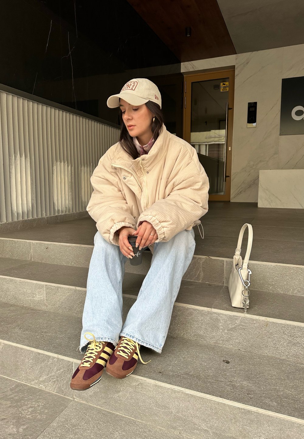 Υπερμεγέθη τζάκετ κοτλέ σε ανοιχτό μπεζ, συνδυασμένο με ανοιχτό μπλε τζιν με φαρδύ πόδι. Καφέ και μπορντό αθλητικά παπούτσια με κίτρινες λεπτομέρειες. Μικρή λευκή τσάντα.