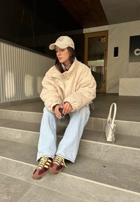 Υπερμεγέθη τζάκετ κοτλέ σε ανοιχτό μπεζ, συνδυασμένο με ανοιχτό μπλε τζιν με φαρδύ πόδι. Καφέ και μπορντό αθλητικά παπούτσια με κίτρινες λεπτομέρειες. Μικρή λευκή τσάντα.