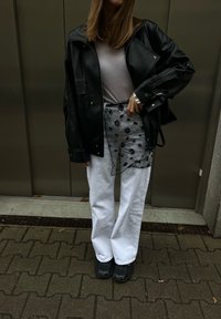 Άτομο που φορεί μαύρο δερμάτινο μπουφάν, διαφανές μαύρο φλοράλ ποδιά πάνω από λευκό παντελόνι και μαύρα παπούτσια, στέκεται σε σκοτεινή πλακόστρωτη επιφάνεια εσωτερικού χώρου.