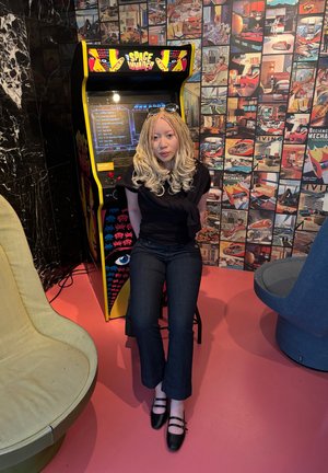 Cabina de juego arcade con un exterior amarillo vibrante que presenta gráficos retro. Persona sentada con ropa oscura, al lado de una silla moderna verde.
