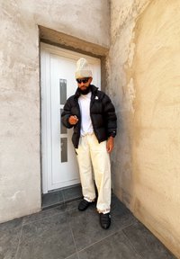 Μαύρο μπουφάν, λευκό T-shirt, παντελόνι σε κρέμα απόχρωση και μαύρα παπούτσια χωρίς κορδόνια. Στοίχειο σε τοίχο με υφή, κρατώντας ένα τηλεκοντρόλ.