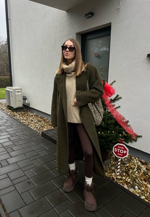 Mujer con abrigo verde oscuro y gafas de sol de pie en una acera mojada cerca de un pequeño árbol de Navidad decorado con un cartel que dice "Santa DETENTE aquí".
