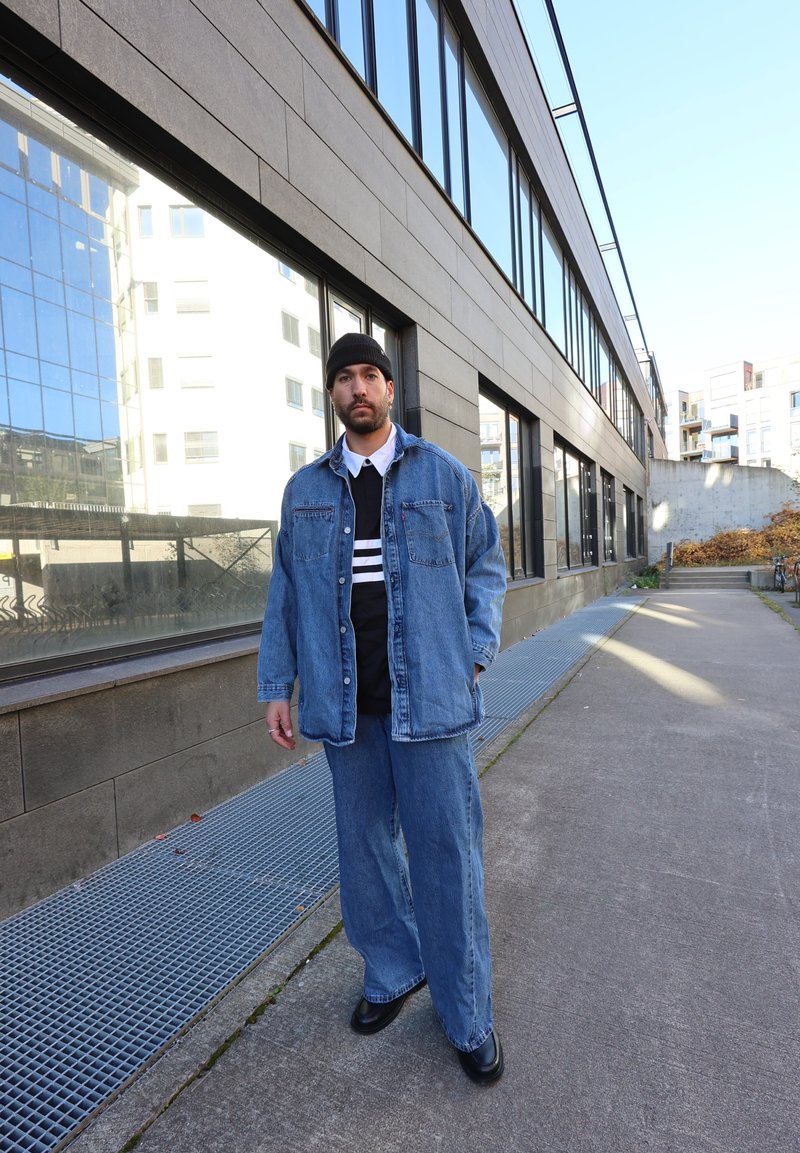 Denim jacket over a black and white striped shirt, paired with wide-leg denim pants and black shoes, against a modern building backdrop.