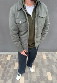 Mees, kes kannab oliivrohelist jope rohelist fliisi peal, tumedad teksad, valged tossud roheliste paeltega, hoiab käes musti päikeseprille, seisab plaaditud tänavakivil.