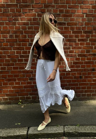Top corto marrón, falda blanca fluida con borde de encaje y bailarinas perforadas de color claro. Modelo con cabello suelto frente a una pared de ladrillo.