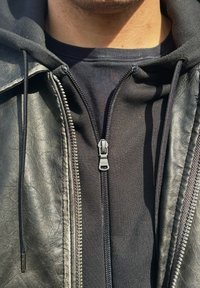 Jaqueta de couro preta com um fecho visível, sobre uma sweat hoodie cinza. O fecho tem um logótipo na aba. Material texturizado notado.