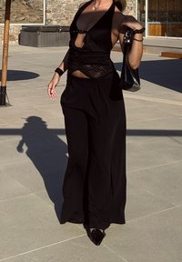 Črni top na vrvico z izrezom in čipkastim detajlom, kombiniran z visokim pasom črnih hlač. Dodatki vključujejo črno torbico in čevlje.