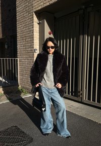 Femme portant un manteau en fourrure noire, un pull gris, un large jean bleu, des lunettes de soleil, et tenant un sac à main noir, debout dans une rue urbaine ensoleillée.