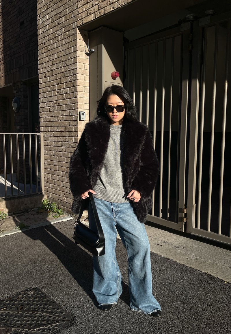 Femme portant un manteau en fourrure noir, un pull gris, un jean large bleu, des lunettes de soleil et tenant un sac à main noir, debout dans une rue urbaine ensoleillée.