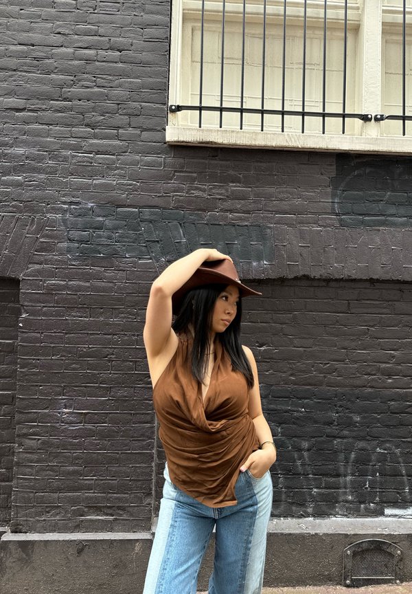 Top marrone drappeggiato abbinato a jeans patchwork blu chiaro e scuro. La modella indossa un cappello marrone, posando davanti a un muro di mattoni scuri e testurizzati.