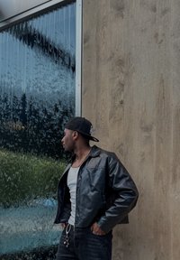 Černá kožená bunda s hladkou texturou, přiléhavým střihem a dlouhými rukávy. V pozadí je vodní prvek a dřevěná stěna.