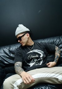 Čierne tričko s potlačou "Favela", svetlosivá čiapka, slnečné okuliare, béžové nohavice s bielymi pruhmi a detailné tetovania na rukách. Čierna kožená pohovka.