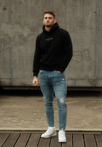 Black hooded sweatshirt with front text, teamed with light blue ripped jeans and white sneakers, set against a concrete backdrop.