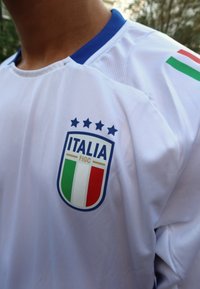 Bílý fotbalový dres s modrým límečkem, znak Itálie s zeleno-bílo-červeným štítem, pět hvězd nad ním. Obsahuje akcenty italské vlajky.