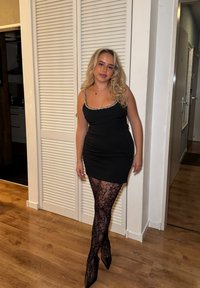 Jeune femme aux cheveux blonds bouclés portant une robe noire à bretelles scintillantes, des collants noirs à motifs et des talons noirs, debout à l'intérieur.