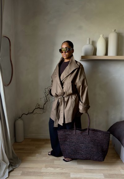 Mujer con chaqueta beige oversize y gafas de sol sostiene un bolso grande de mimbre, de pie en una habitación minimalista con decoración neutra y suelo de madera.