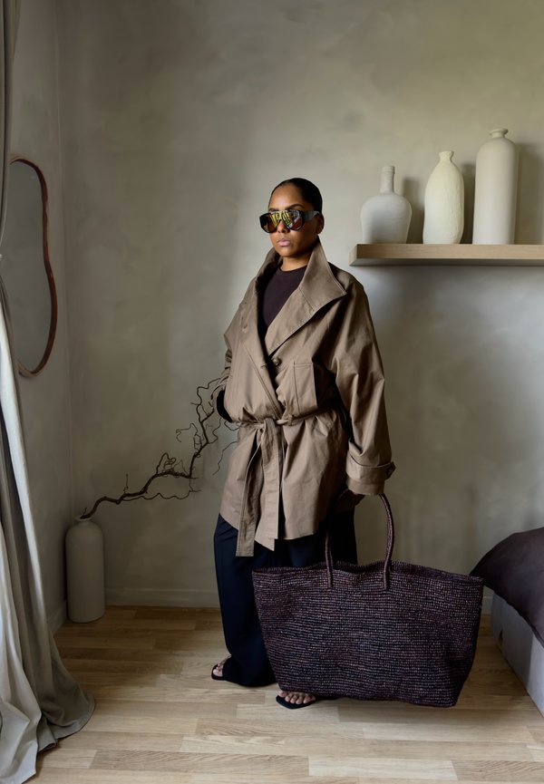 Frau in übergroßer beiger Jacke und Sonnenbrille hält eine große geflochtene Tasche, steht in einem minimalistischen Raum mit neutraler Einrichtung und Holzboden.