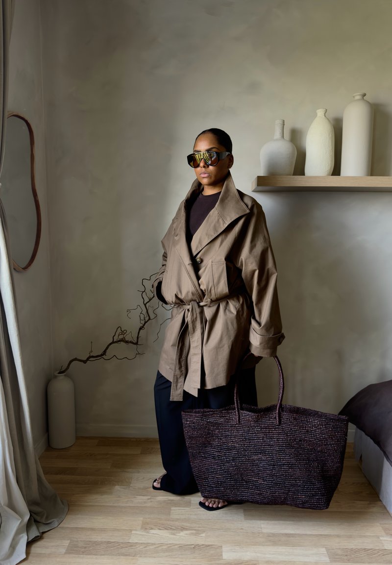 Kvinne i stor beige jakke og solbriller holder en stor vevd veske, står i et minimalistisk rom med nøytral innredning og tregulv.