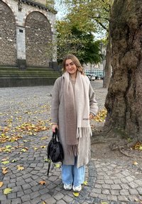 Abrigo gris claro con una bufanda larga en degradado beige y marrón, jeans azules casuales y zapatillas blancas, sosteniendo un bolso negro sobre suelo adoquinado.