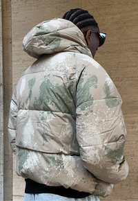 Puf jakna s bež i zelenim uzorkom prskanja boje, s kapuljačom i elastičnim manžetama, prikazana sa stražnje strane na neutralnoj pozadini.