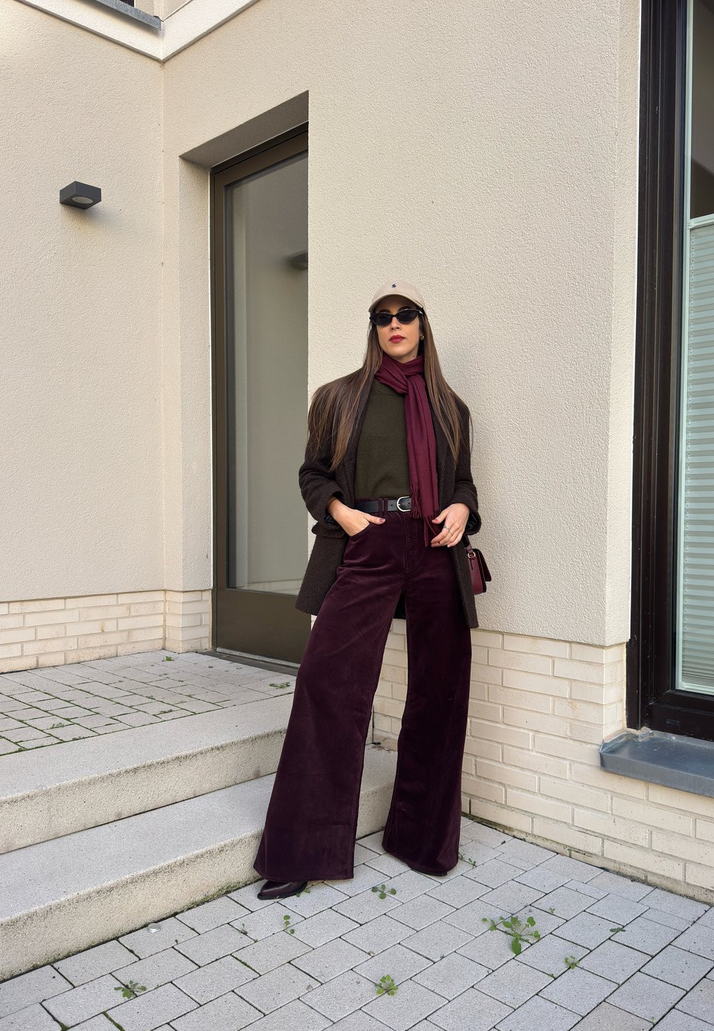 Suurreiset tummanpunaiset corduroy-housut, oliivinvihreä paita, tummanruskea bleiseri, maroonihuivi, beige hattu ja mustat aurinkolasit neutraalia seinää vasten.