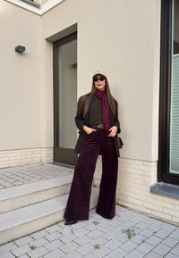 Suurreiset tummanpunaiset corduroy-housut, oliivinvihreä paita, tummanruskea bleiseri, maroonihuivi, beige hattu ja mustat aurinkolasit neutraalia seinää vasten.