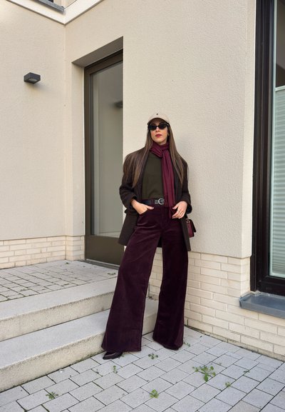 Pantalones de pana burdeos de pierna ancha, blusa verde oliva, blazer marrón oscuro, bufanda marrón, gorra beige y gafas de sol negras contra una pared neutral.