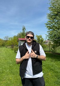 Schwarze ärmellose Weste über einem weißen Shirt, mit Reißverschluss und Logo-Patch. Kurzes dunkles Haar, Sonnenbrille und sichtbare Tattoos. Außeneinstellung.