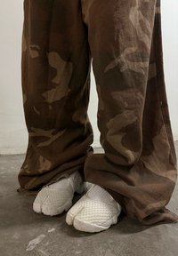 Persoon in bruine camouflagebroeken en witte gespleten-teenschoenen staat op een betonnen vloer naast een witte muur.