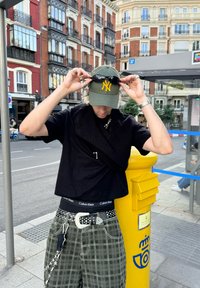 Fekete, rövid ujjú póló egy textúrált pulóverréteggel, zöld sapka sárga logóval, kockás nadrág, kiegészítő láncok és napszemüveg. Sárga oszlop a háttérben.