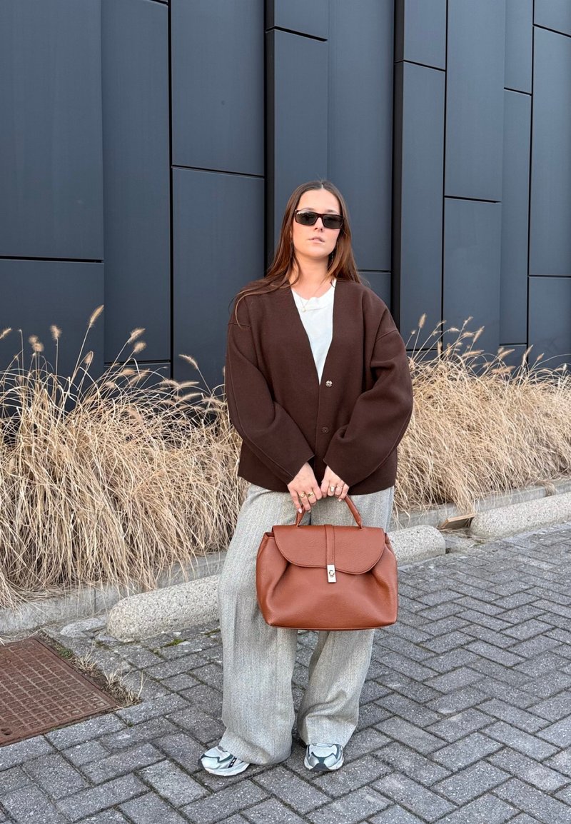 Jeune femme portant des lunettes de soleil, une veste marron oversize, un pantalon large gris, des baskets blanches, tenant un grand sac à main marron, debout sur une rue pavée.