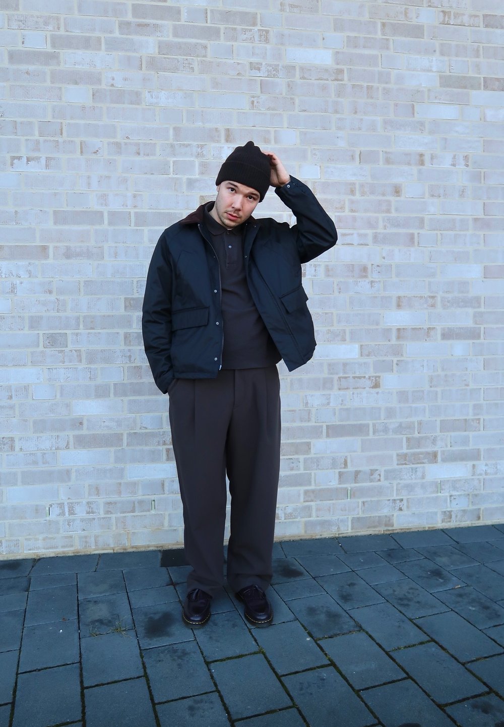 Must must, pruun krae, tume sviiter ja pruunid püksid. Pruun beanie, seistes hallil sillutisel tekstureeritud tellisseina vastu.