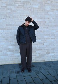 Must must, pruun krae, tume sviiter ja pruunid püksid. Pruun beanie, seistes hallil sillutisel tekstureeritud tellisseina vastu.