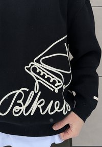 Personne portant un pull noir avec un motif brodé blanc représentant une chaussure de sport et un texte en écriture cursive, main reposant près de la taille sur une chemise blanche.