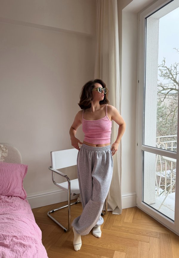 Ženska z očali za sonce, roza majico brez rokavov in sivimi trenirkami stoji ob belem stolu ob oknu z zaveso v spalnici.