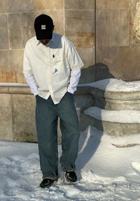 Homme portant une casquette noire, des chemises blanches superposées avec une tache, un jean ample et des chaussures noires, debout sur la neige à côté d'un mur en pierre avec des détails sculptés.