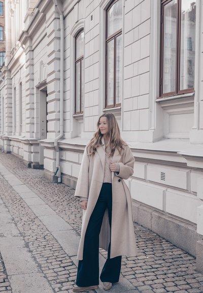 Mujer con abrigo largo beige y pantalones negros de pie en una calle adoquinada junto a un edificio blanco con grandes ventanas.