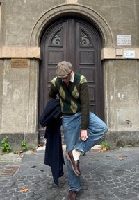Pull en tricot avec un motif argyle vert et marron, jean en denim, chaussures marron, chaussettes claires et casquette plates ; debout près des portes en bois.