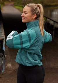 Türkisfarbener Windbreaker mit Streifenakzenten, hohem Kragen und Reißverschlusstaschen. Wird über schwarzen Leggings getragen und besteht aus leichtem Material.