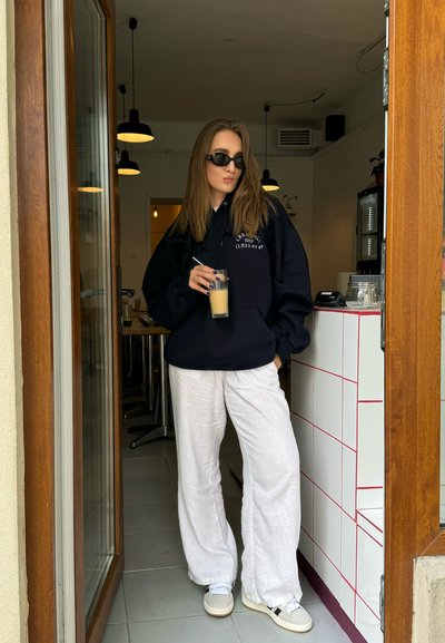 Sudadera negra de gran tamaño con texto blanco, pantalones de lino blancos y zapatillas beige. Sosteniendo un vaso de café helado, de pie junto a una puerta.