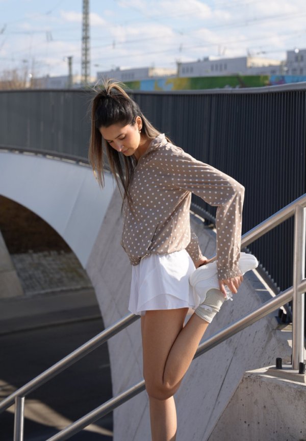 Νεαρή γυναίκα με πουά μπλούζα και λευκή φούστα τεντώνει το πόδι κρατώντας το πόδι της στη ράγα των εξωτερικών σκαλοπατιών.