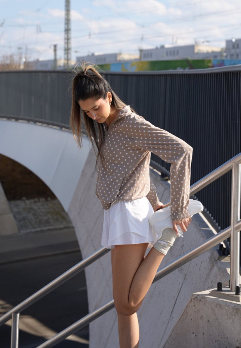 Νεαρή γυναίκα με πουά μπλούζα και λευκή φούστα τεντώνει το πόδι κρατώντας το πόδι της στη ράγα των εξωτερικών σκαλοπατιών.