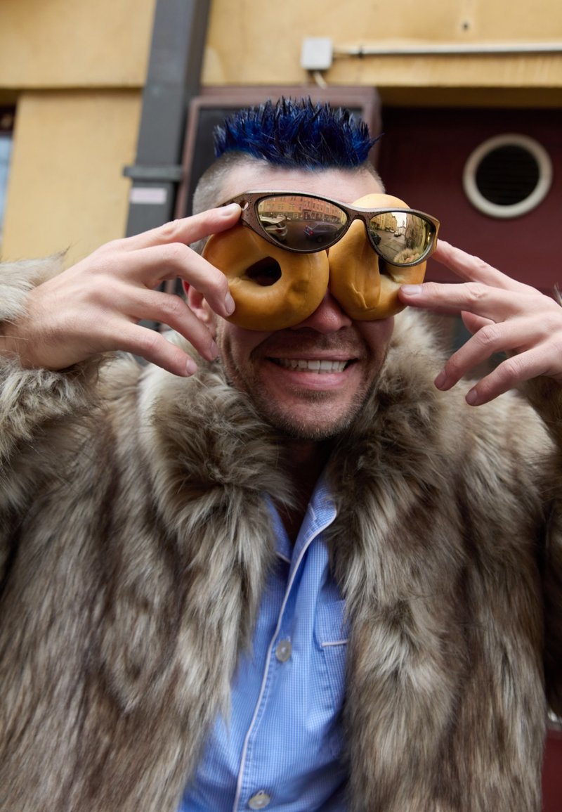 Manteau en fausse fourrure marron clair, lunettes de soleil bleues et lunettes en forme de beignet tenues à la main, sur un fond neutre.