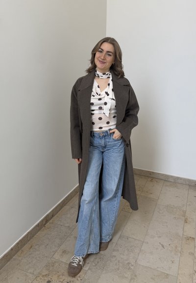 Mujer sonriendo, vestida con un abrigo largo oscuro sobre una blusa blanca con lunares, vaqueros de pierna ancha azul claro y zapatillas marrones, de pie en un interior.