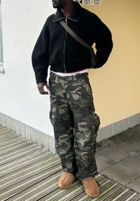 Sweater preto com zip e calças camufladas largas com bolsos, e chinelos castanhos. De pé sobre uma superfície de madeira e azulejo, fundo neutro.