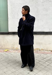 Abrigo de lana azul oscuro con cuello alto, combinado con pantalones anchos negros y zapatos negros, posando frente a una pared blanca texturizada.