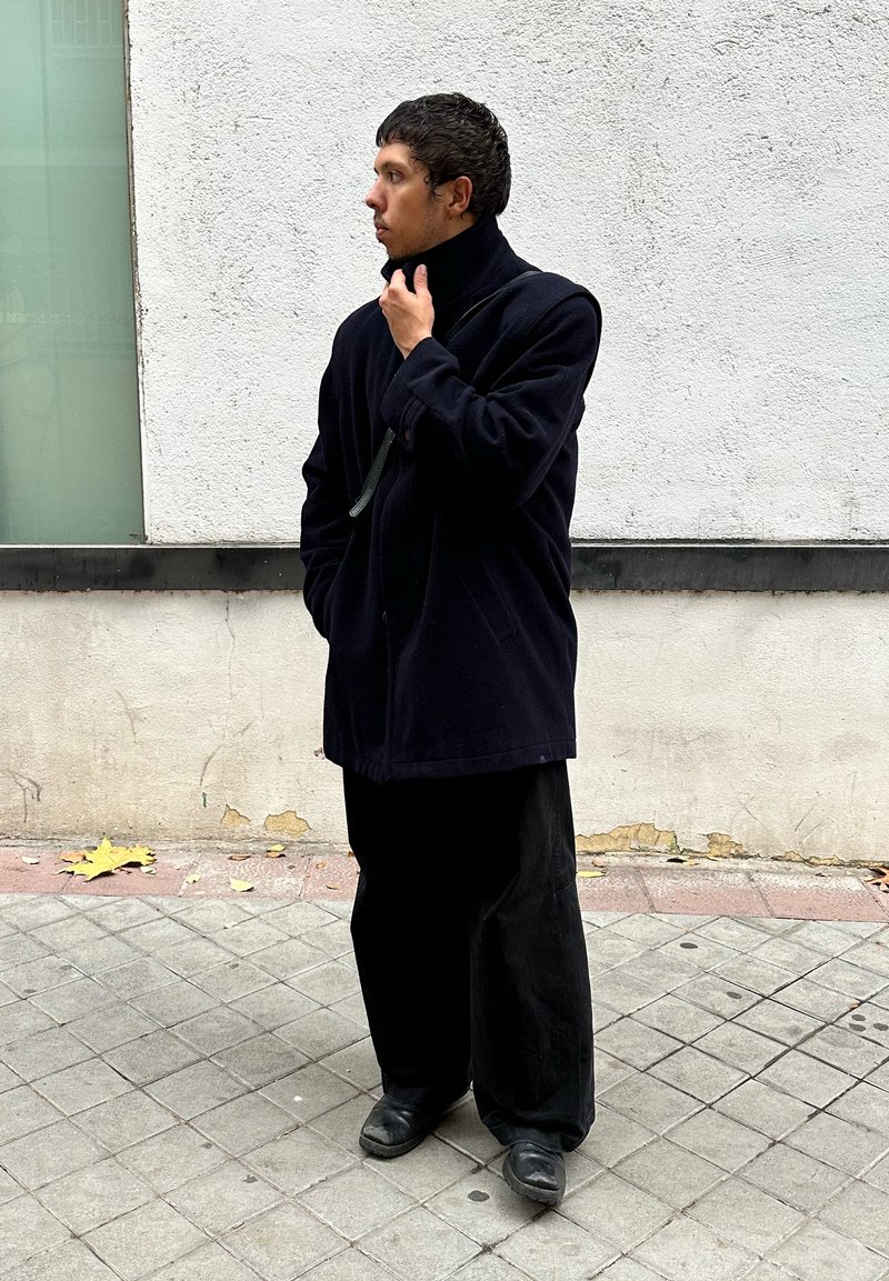 Dark navy wool coat with a high collar, paired with wide-legged black trousers and black shoes, standing against a textured white wall.