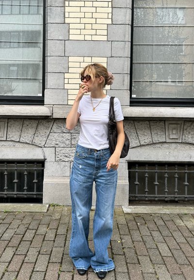 Modelo vistiendo una camiseta blanca ajustada, jeans azules de cintura alta con un ajuste relajado, bolso negro y gafas de sol de gran tamaño, de pie sobre un pavimento de ladrillos.